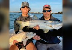 A teenage angler and a man in sunglasses holding a big snook and a redfish on a boat