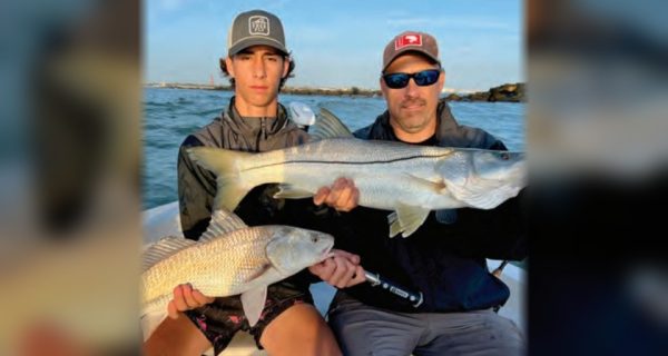 A teenage angler and a man in sunglasses holding a big snook and a redfish on a boat