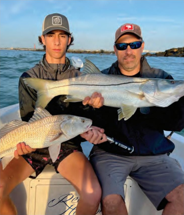A teenage angler and a man in sunglasses holding a big snook and a redfish on a boat