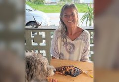 A smiling woman holding a smoked fish on a wooden board as a shaggy dog sniffs it