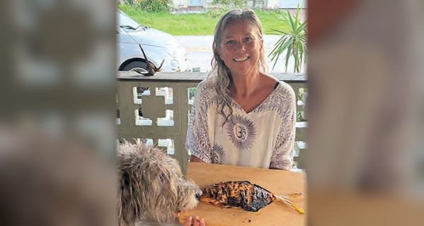 A smiling woman holding a smoked fish on a wooden board as a shaggy dog sniffs it