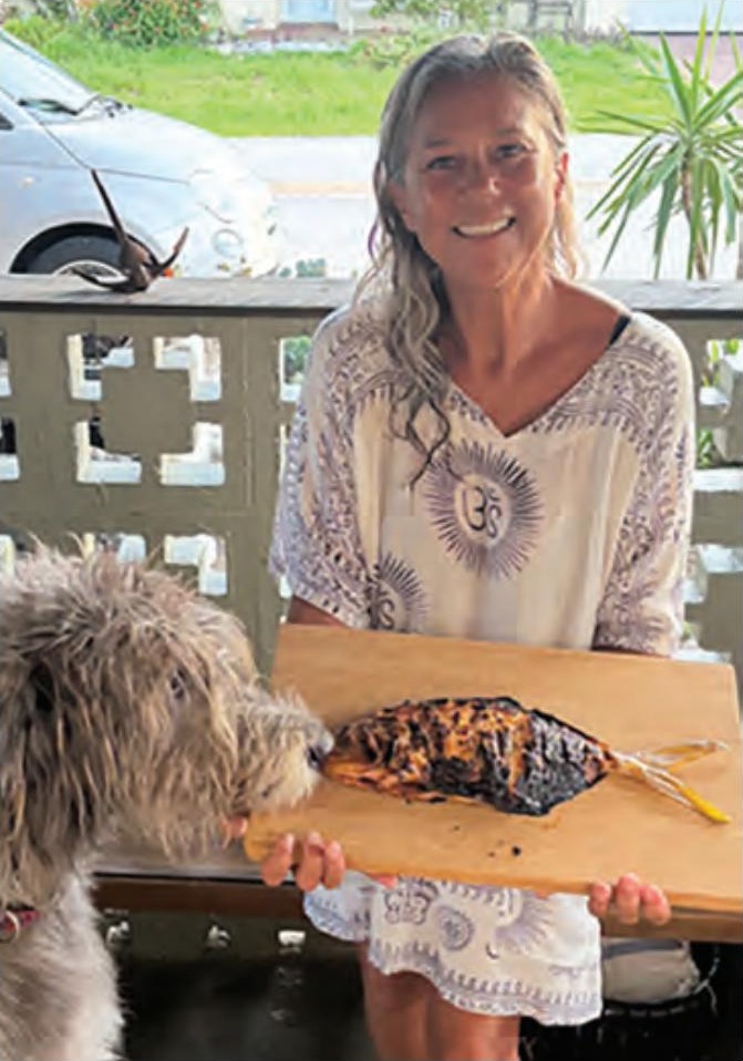 A smiling woman holding a smoked fish on a wooden board as a shaggy dog sniffs it