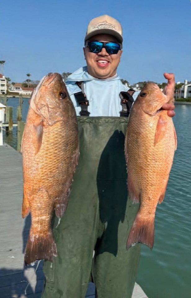 A smiling angler in waders and blue sunglasses holding up two big snapper on a dock