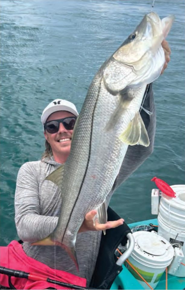 An angler in a white cap and sunglasses holding up a big snook in his kayak