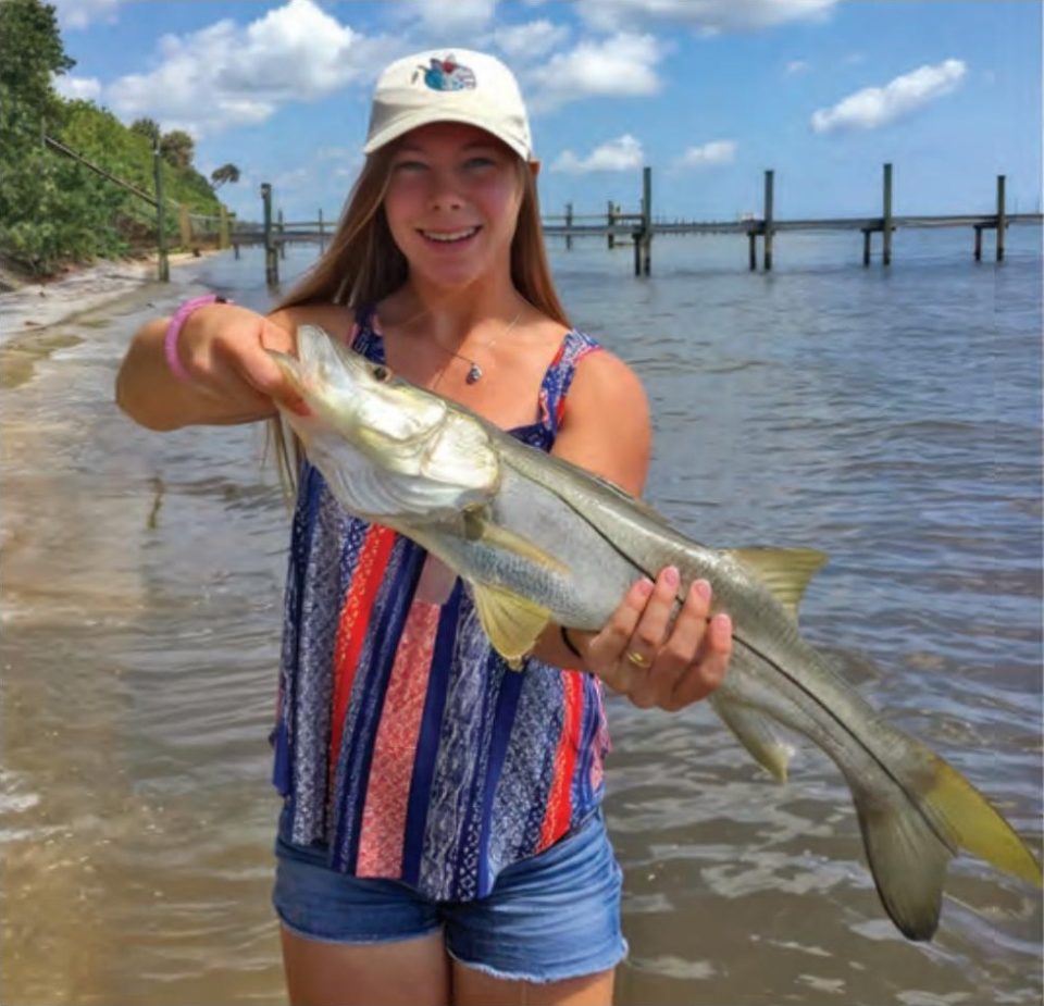 A smiling young woman in a cap holding a snook while wading along a sandy shoreline with a pier behind her