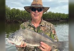 An angler in a straw hat and camouflage shirt holding a black drum in front of the mangroves