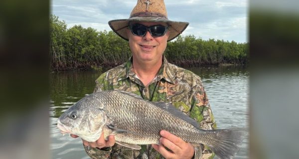 An angler in a straw hat and camouflage shirt holding a black drum in front of the mangroves