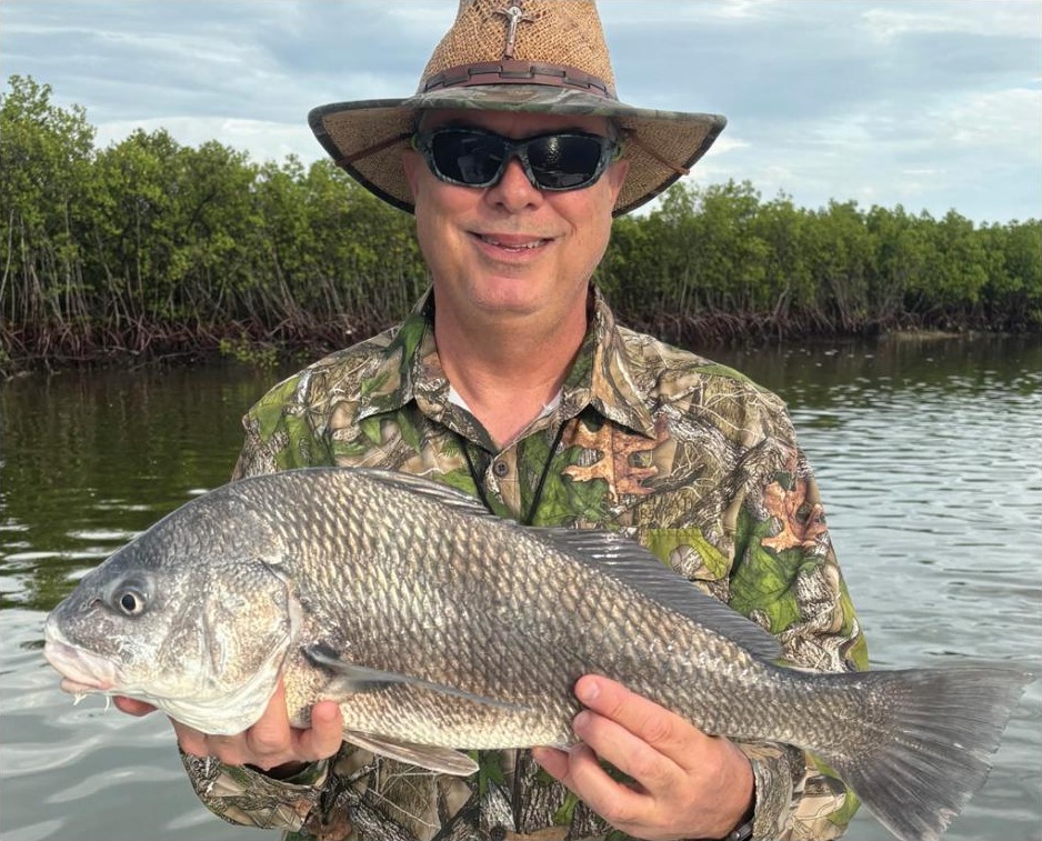An angler in a straw hat and camouflage shirt holding a black drum in front of the mangroves