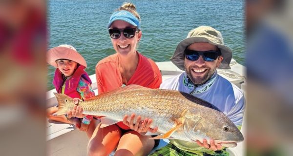 A young girl in a pink sun hat, a woman in a visor, and a man in a bucket hat holding a big redfish on a boat