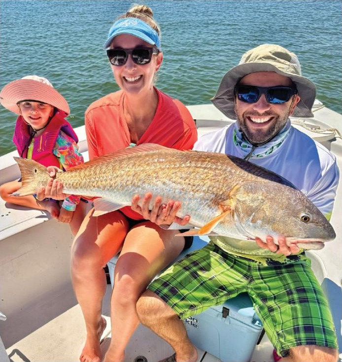 A young girl in a pink sun hat, a woman in a visor, and a man in a bucket hat holding a big redfish on a boat