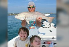 An angler in sunglasses holding up a redfish on a boat, with two smiling boys in front of him and a lighthouse in the distance
