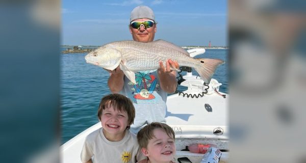 An angler in sunglasses holding up a redfish on a boat, with two smiling boys in front of him and a lighthouse in the distance