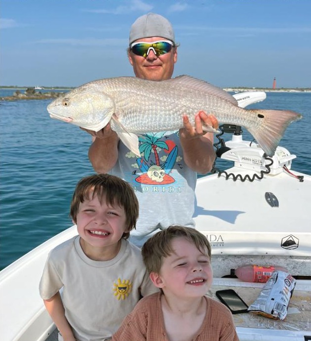 An angler in sunglasses holding up a redfish on a boat, with two smiling boys in front of him and a lighthouse in the distance
