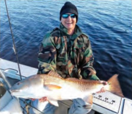 An angler in a camouflage jacket and knit cap holding a big redfish on a boat