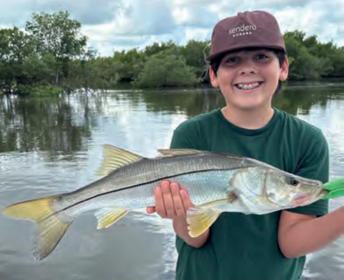 A smiling boy in a cap holding a snook in front of the mangroves