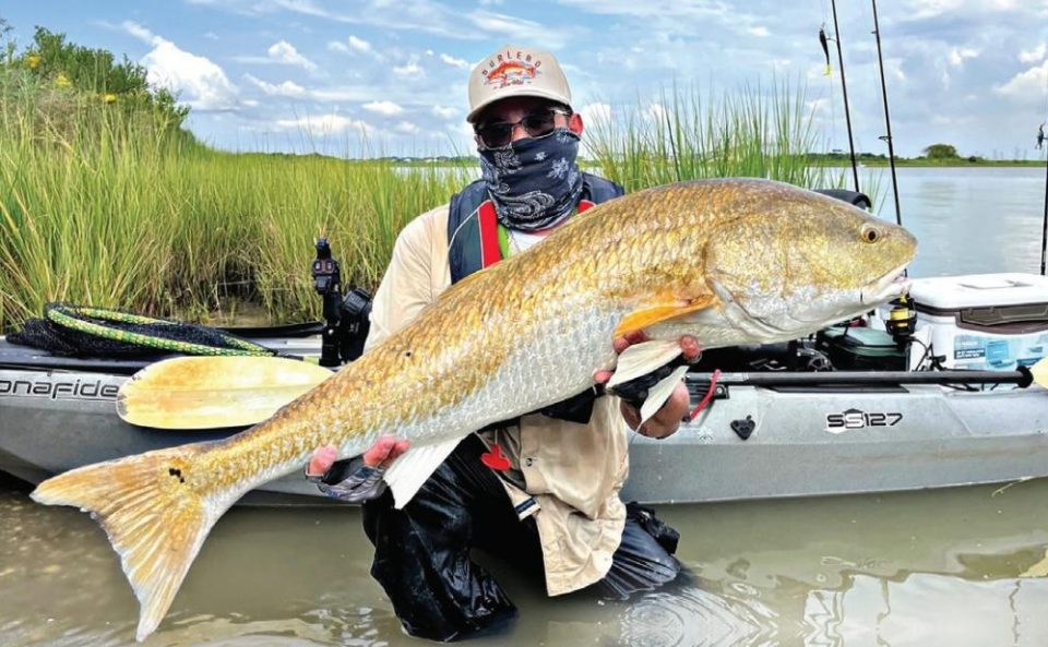 An angler in a face gaiter kneeling in the shallows beside a kayak with a big redfish