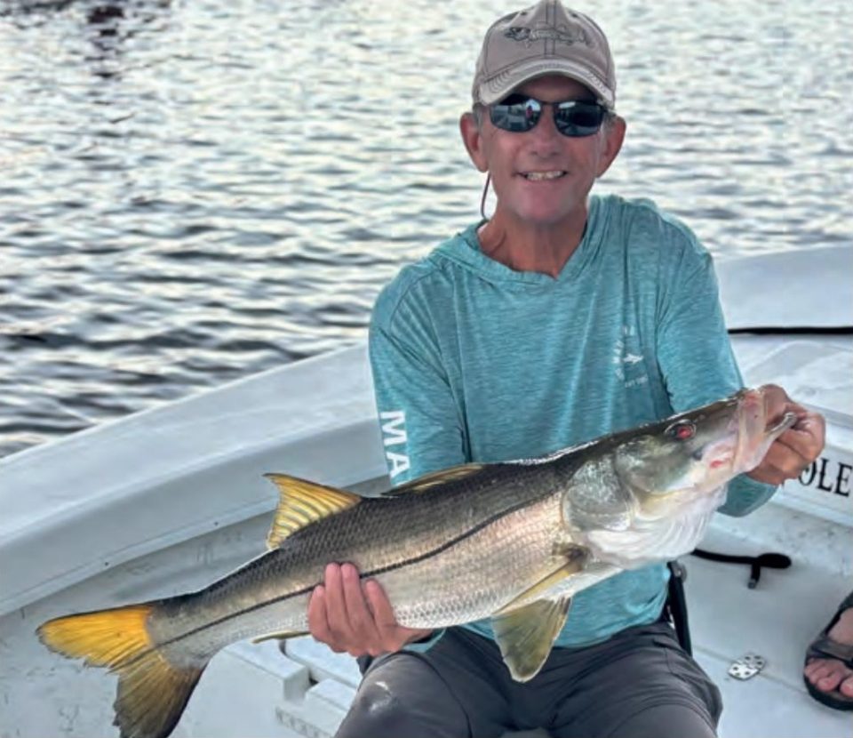 An angler in a cap and sunglasses holding a big snook on a boat