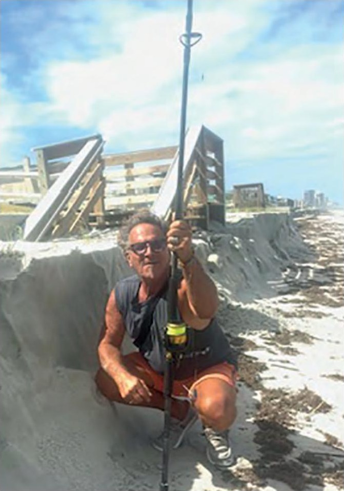 Marco Pompano crouching with a surf rod below a sand cliff cut into the beach at Wilbur by the Sea, with a damaged beach walkover behind him