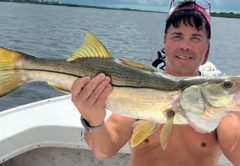 A shirtless angler in a cap holding a big snook on a boat