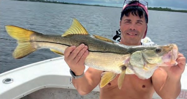 A shirtless angler in a cap holding a big snook on a boat