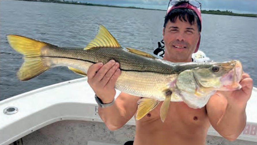 A shirtless angler in a cap holding a big snook on a boat