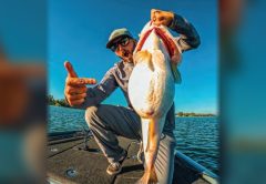 An angler pointing at a big bass he holds up by the jaw on the deck of a bass boat