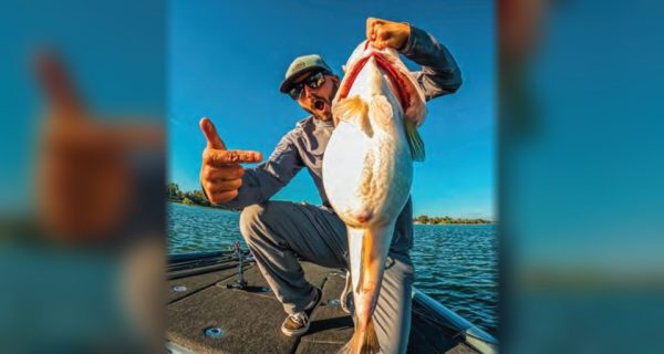 An angler pointing at a big bass he holds up by the jaw on the deck of a bass boat