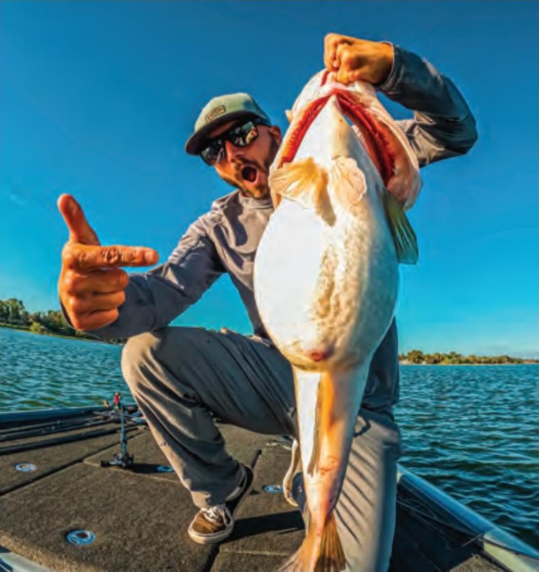 An angler pointing at a big bass he holds up by the jaw on the deck of a bass boat