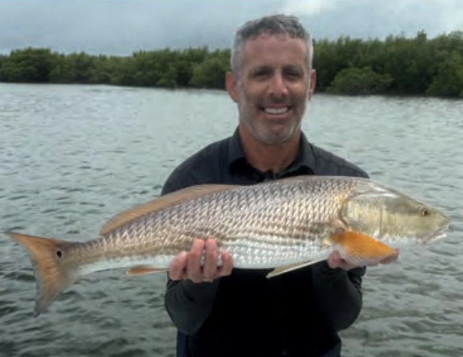 A smiling angler holding a redfish in front of the mangroves