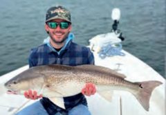 An angler in a camouflage cap and plaid shirt holding a big redfish on the bow of a boat