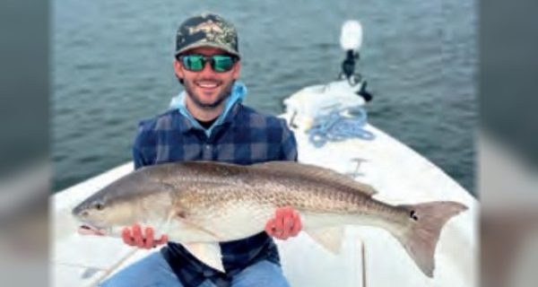 An angler in a camouflage cap and plaid shirt holding a big redfish on the bow of a boat