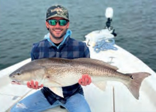 An angler in a camouflage cap and plaid shirt holding a big redfish on the bow of a boat