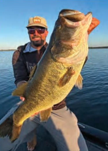 An angler in a cap and sunglasses holding a big largemouth bass on a boat