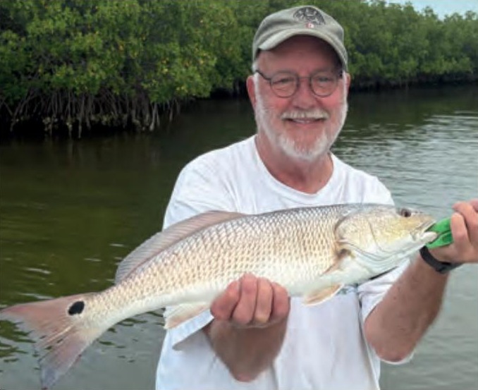 An angler in a cap and glasses holding a redfish in front of the mangroves