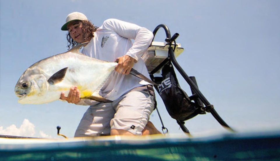 An angler kneeling on a stand-up paddleboard holding a permit