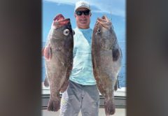 An angler in a white cap and sunglasses holding two big grouper offshore