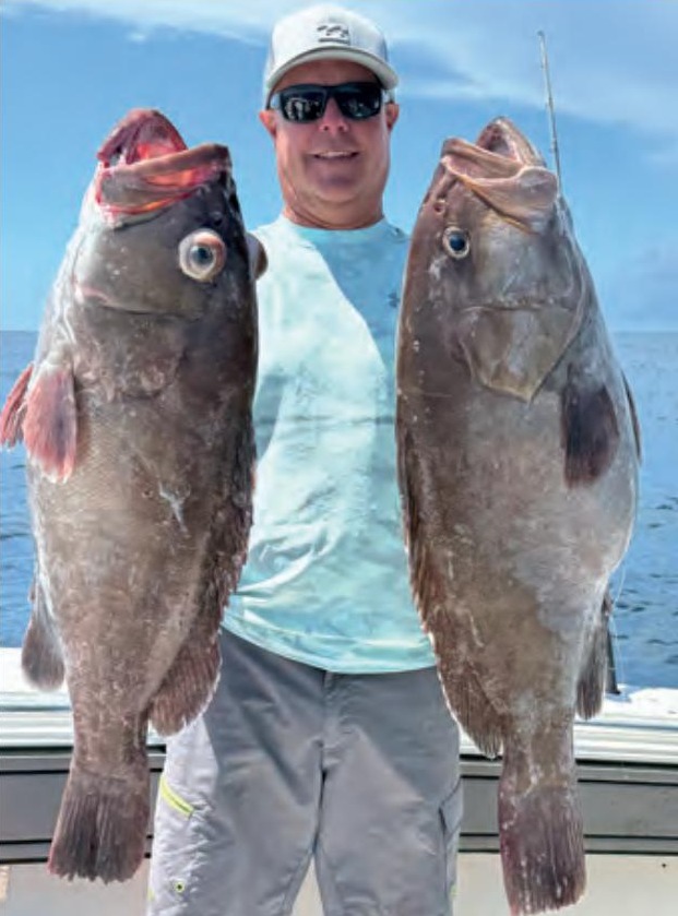 An angler in a white cap and sunglasses holding two big grouper offshore