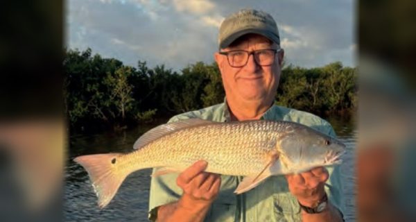 An angler in glasses and a cap holding a redfish in front of the mangroves