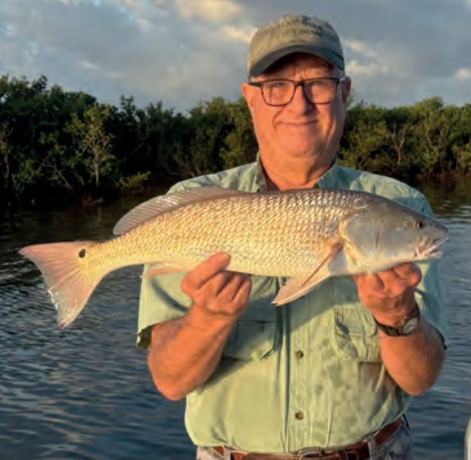 An angler in glasses and a cap holding a redfish in front of the mangroves