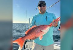 An angler in a cap and sunglasses holding a red snapper on a boat offshore