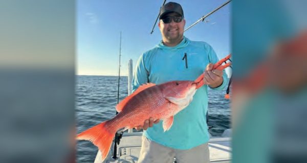 An angler in a cap and sunglasses holding a red snapper on a boat offshore