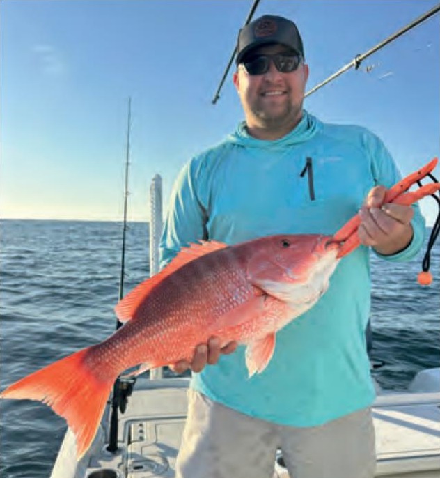 An angler in a cap and sunglasses holding a red snapper on a boat offshore
