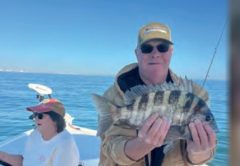 An angler in a tan jacket holding a sheepshead on a boat, with a woman at the helm behind him