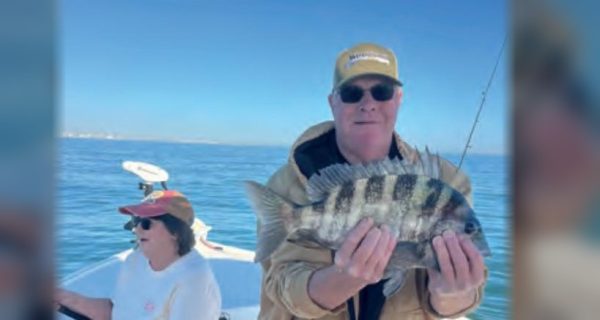 An angler in a tan jacket holding a sheepshead on a boat, with a woman at the helm behind him
