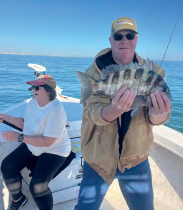 An angler in a tan jacket holding a sheepshead on a boat, with a woman at the helm behind him