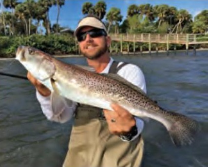 A wading angler in a visor and sunglasses holding a big speckled trout