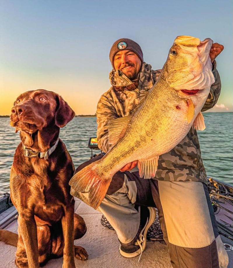 An angler holding a giant largemouth bass next to a chocolate Lab on a boat