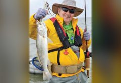 An angler in a sun hat and yellow jacket holding a speckled trout on the flats