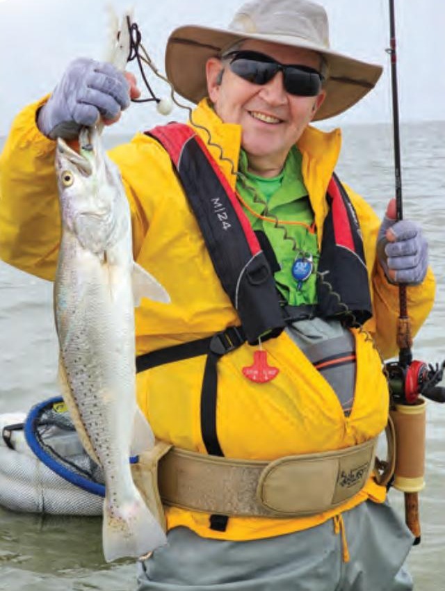 An angler in a sun hat and yellow jacket holding a speckled trout on the flats