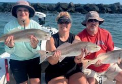 Three anglers each holding a redfish on a boat in front of the jetty rocks